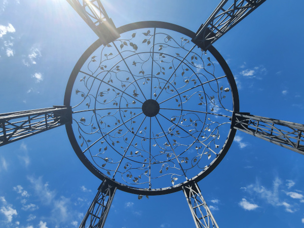 Low-angle view looking up at the intricate, ornamental wrought-iron dome of a garden gazebo featuring leaf-pattern scrollwork against a bright blue sky.